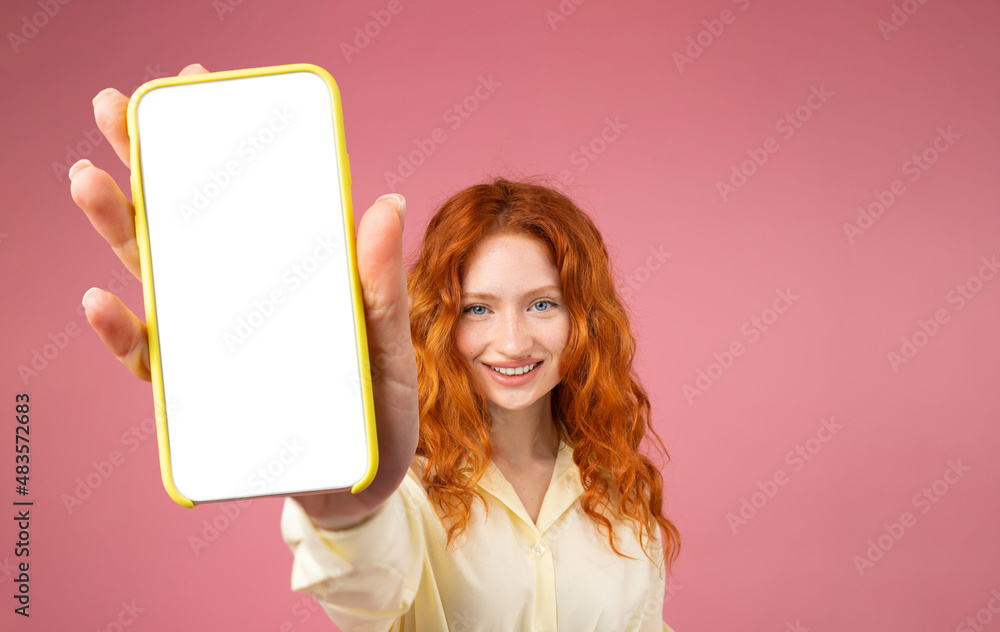 Portrait of excited redhead girl holding big smartphone with white blank screen in hand, showing close to camera. Gadget with empty free space for mock up, banner isolated on pink background.