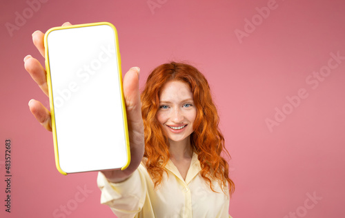 Portrait of excited redhead girl holding big smartphone with white blank screen in hand, showing close to camera. Gadget with empty free space for mock up, banner isolated on pink background.