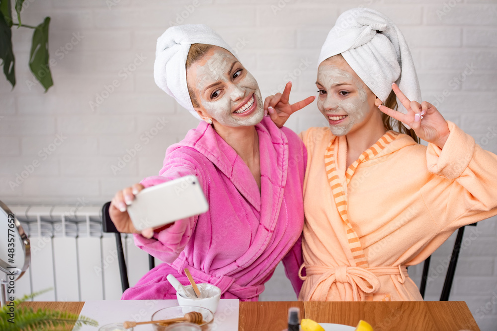 Mother and her daughter taking selfie by smartphone with facial mask on, bathrobe and towel on head Mother and her daughter taking selfie by smartphone with facial mask on, bathrobe and towel on head