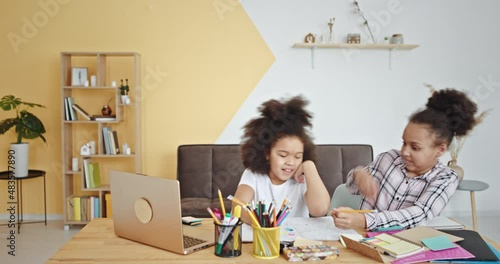 Smiling mixed race junior schoolgirls with kinky hair draw on paper with colorful pencils near laptop at table in children room photo