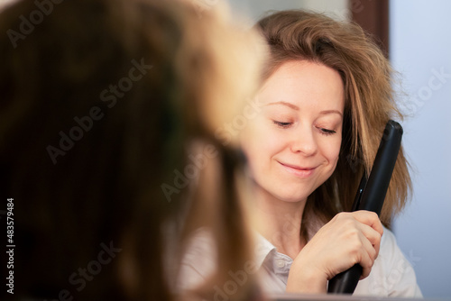 woman straightens her hair with an iron in front of a mirror