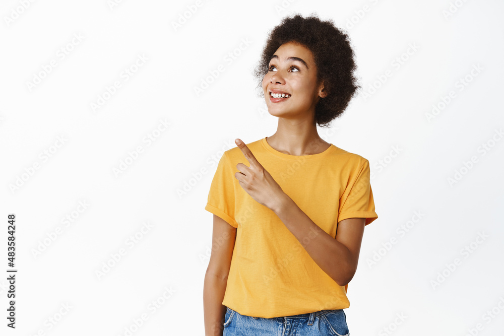 Smiling black girl pointing and looking at upper left corner with pleased carefree smile, reading promo text, showing company brand, logo, white background Smiling black girl pointing and looking at upper left corner with pleased carefree smile, reading promo text, showing company brand, logo, white background
