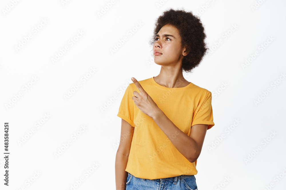 Confused black female model pointing, looking at upper left corner logo, frowning perplexed by advertisement, standing over white background Confused black female model pointing, looking at upper left corner logo, frowning perplexed by advertisement, standing over white background