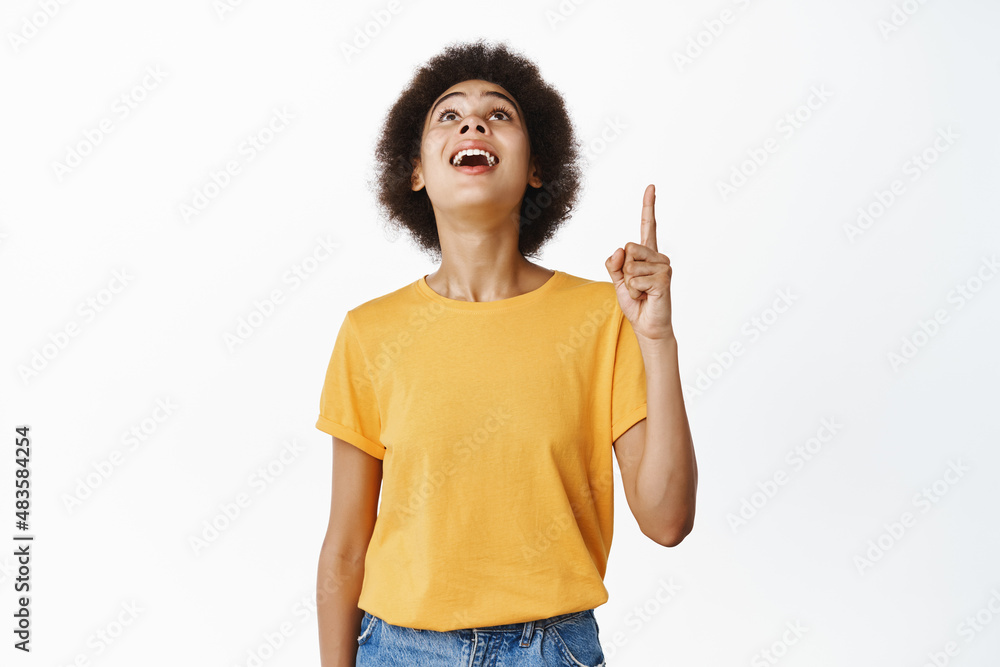 Image of excited smiling black girl checking out smth cool, pointing finger and looking up with amazed excitement, standing over white background Image of excited smiling black girl checking out smth cool, pointing finger and looking up with amazed excitement, standing over white background