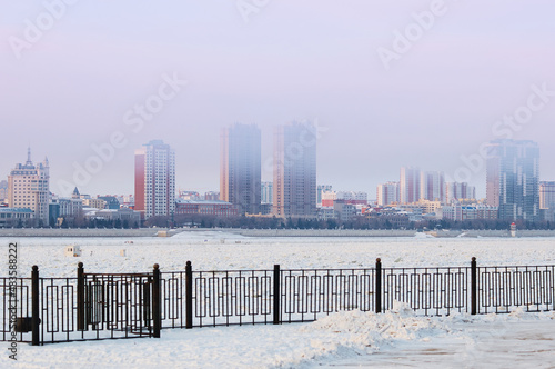 Frosty haze in winter over the border river Amur. View of the city of Heihe, China from the embankment of the city of Blagoveshchensk, Russia. Tall buildings behind a small fence. © Алексей Игнатов