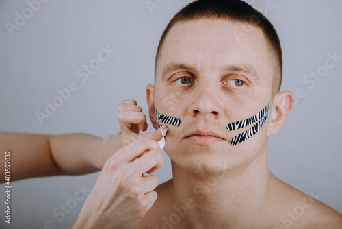 Portrait of a guy with a plaster on his face. Kinesio taping. The girl's hands glue a striped patch on the guy's face.