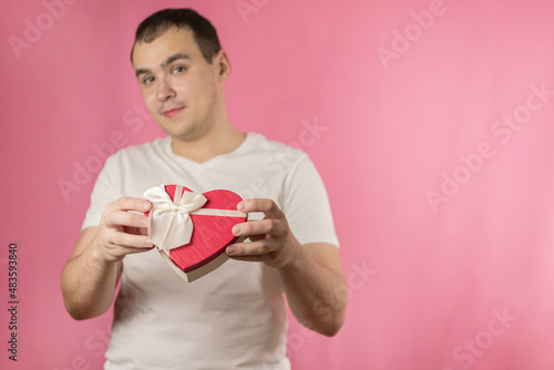 A young man in a white T-shirt on a pink background with a gift for Valentine's day. A man with a gift on a pink background. The man is out of focus, the focus is on the gift.
