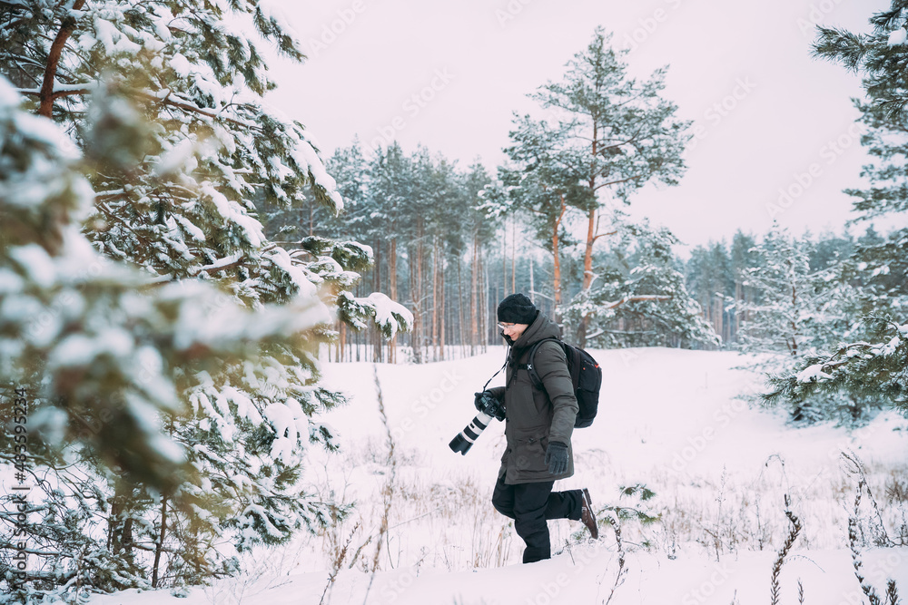 Young Happy Man Backpacker With Photo Camera Taking Photo In Winter Snowy Forest. Active Hobby. Hiker Walking In Snowy Pinewood Forest.
