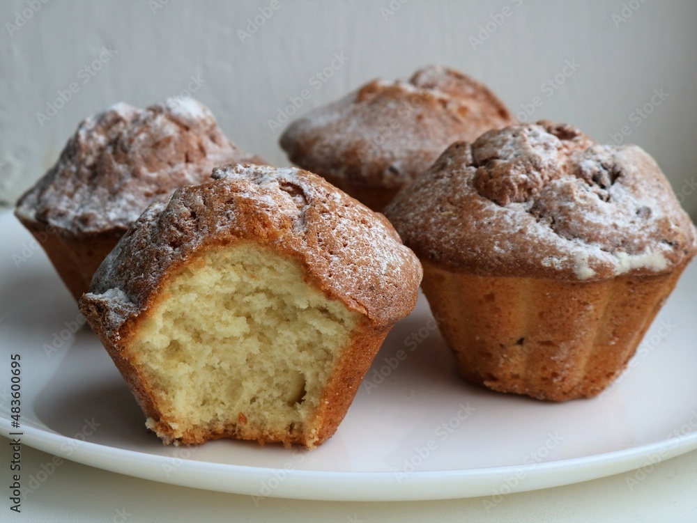appetizing bitten cupcake against the background of three whole muffins sprinkled with powdered sugar standing on a white plate against a light texture wall, a sweet dessert for tea drinking