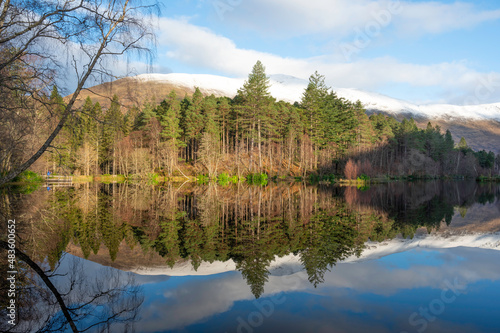 Glencoe lochan in Scottish Highlands in winter. Mirror reflection of trees, blue sky and forest in water. No people.