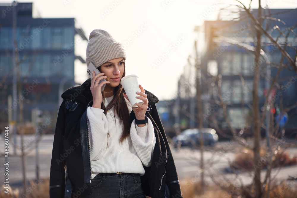 Smiling woman with smart phone and coffe to go standing outdoors and speaking. Modern lifestyle concept.