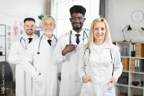 Group of confident practicing doctors in white lab coats and eyeglasses standing together at hospital room. International conference and cooperation concept.