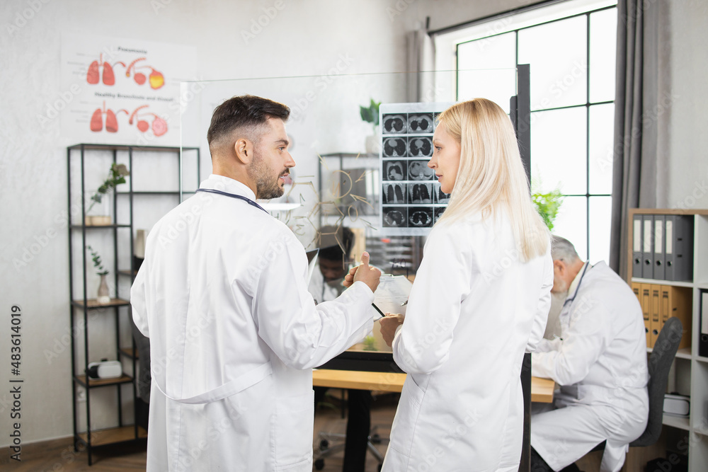 Obraz premium Caucasian male and female medical scientists in white lab coats having discussion while standing near glass flip chart at hospital room. Researchers, cooperation and brainstorming concept.