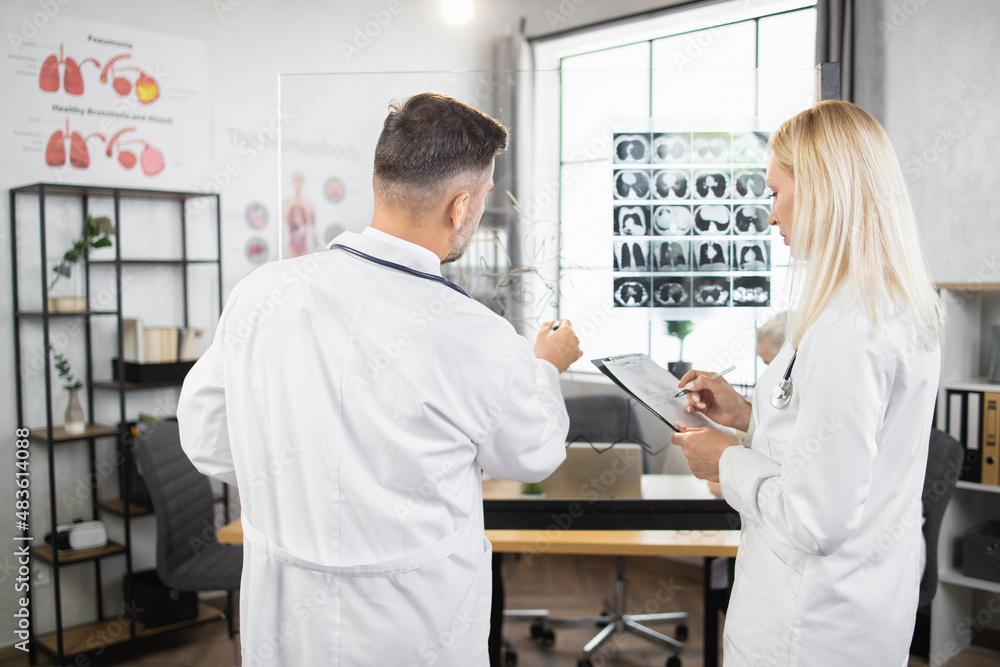 Medical experts in white lab coats writing on flipchart while analysing results of x ray scan. Man and woman having meeting at hospital about severe patient diagnosis. Medical experts in white lab coats writing on flipchart while analysing results of x ray scan. Man and woman having meeting at hospital about severe patient diagnosis.