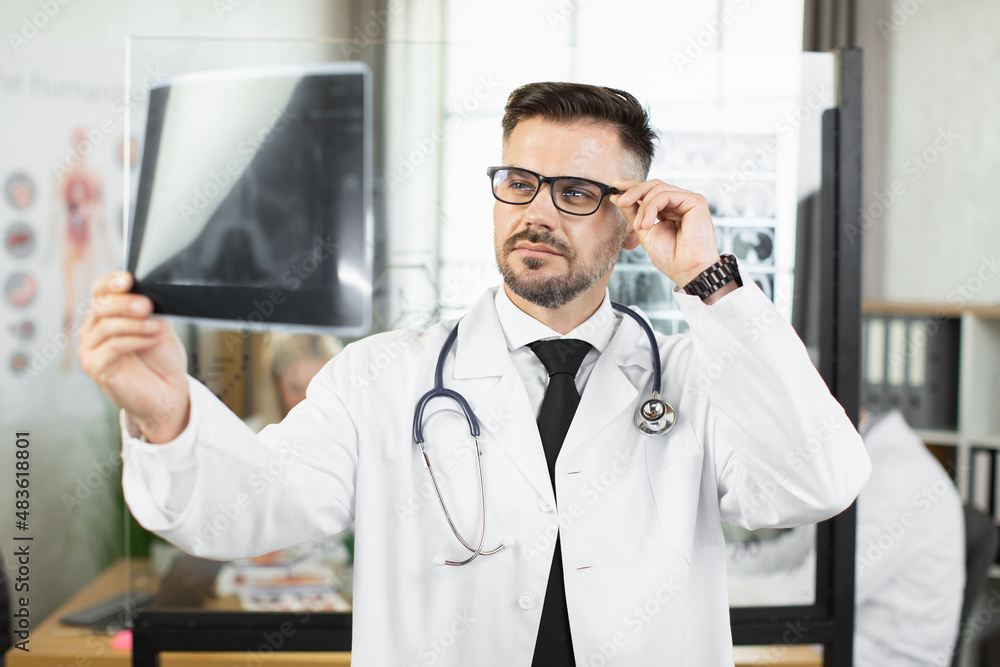 Caucasian male physician in lab coat and eyeglasses examining x ray scan that holding in hands. Qualified doctor during working process at hospital.