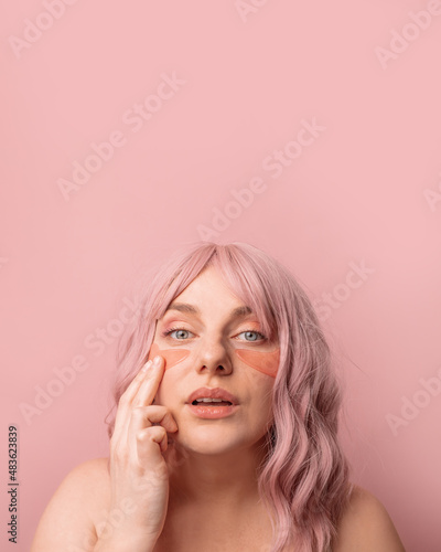 Beautiful woman applying pink under eye patches for dark-circles and puffiness looking at the camera isolated over pink background. Spa, skincare