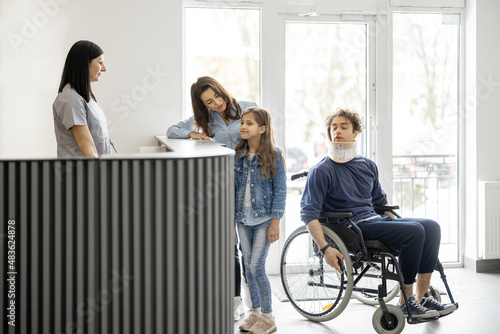 Young people in the lobby of the clinic. Woman with girl talking at the reception desk and medical worker taking care of patient in wheelchair