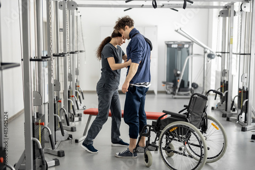 Rehabilitation specialist helps a guy stand out of a wheelchair at rehabilitation center. Concept of physical therapy and support for people with disabilities photo