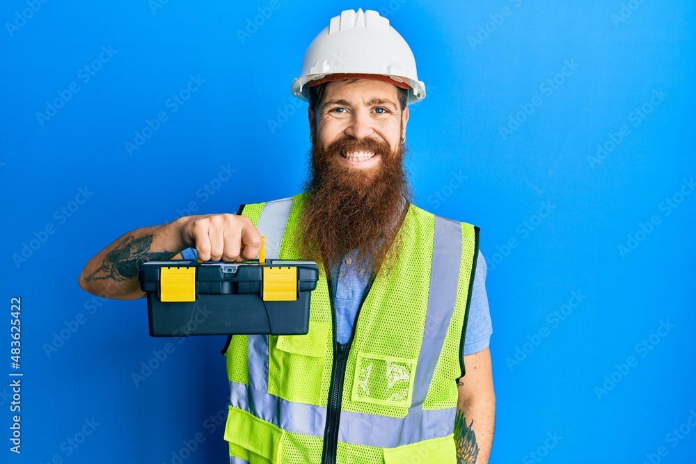 Redhead man with long beard wearing safety helmet and reflective jacket holding toolbox looking positive and happy standing and smiling with a confident smile showing teeth