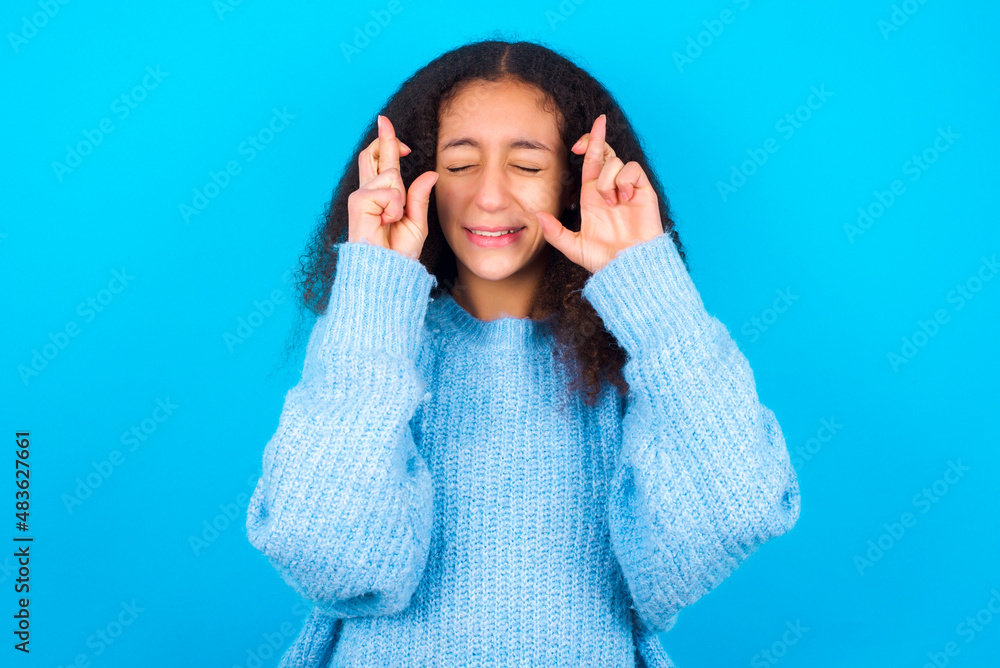 Joyful African teenager girl wearing blue sweater over blue background clenches teeth, raises fingers crossed, makes desirable wish, waits for good news, I have to win. Joyful African teenager girl wearing blue sweater over blue background clenches teeth, raises fingers crossed, makes desirable wish, waits for good news, I have to win.