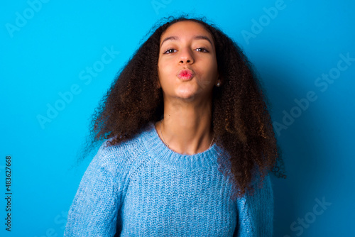 African teenager girl wearing blue sweater over blue background, keeps lips as going to kiss someone, has glad expression, grimace face. Standing indoors. Beauty concept.