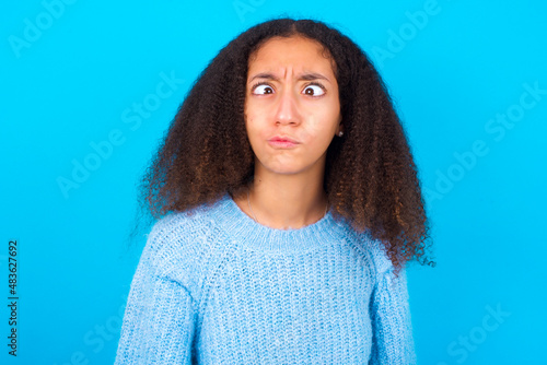 African teenager girl wearing blue sweater over blue background crosses eyes, puts lips, makes grimace with awkward expression has fun alone, plays fool.