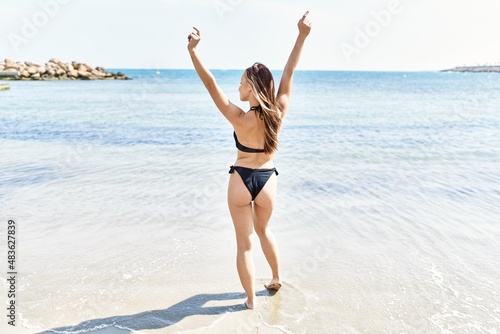 Young cuacasian girl on back view wearing bikini with hands raised up at the beach.