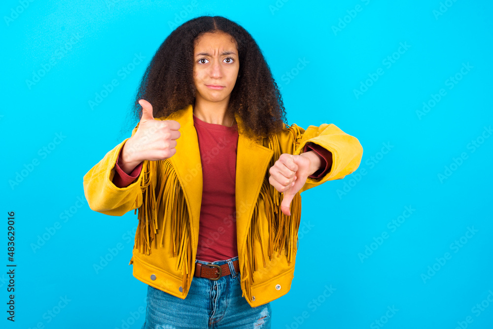 African teenager girl wearing yellow jacket over blue background feeling unsure making good bad sign. Displeased and unimpressed. African teenager girl wearing yellow jacket over blue background feeling unsure making good bad sign. Displeased and unimpressed.