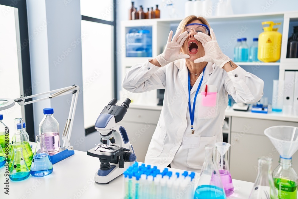 Middle age blonde woman working at scientist laboratory shouting angry out loud with hands over mouth Middle age blonde woman working at scientist laboratory shouting angry out loud with hands over mouth