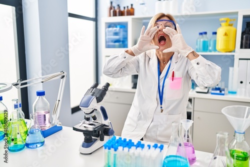 Middle age blonde woman working at scientist laboratory shouting angry out loud with hands over mouth