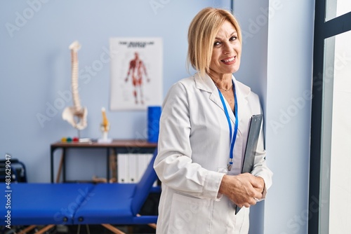 Middle age blonde woman wearing physiotherapist uniform standing at physiotherapy clinic