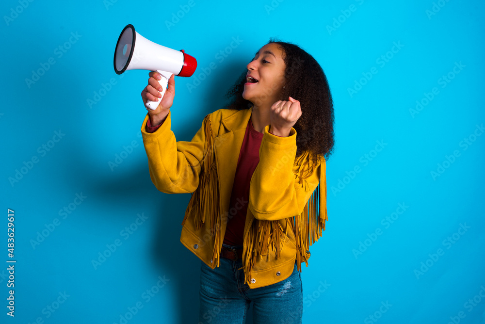 African teenager girl wearing yellow jacket over blue background communicates shouting loud holding a megaphone, expressing success and positive concept, idea for marketing or sales. African teenager girl wearing yellow jacket over blue background communicates shouting loud holding a megaphone, expressing success and positive concept, idea for marketing or sales.