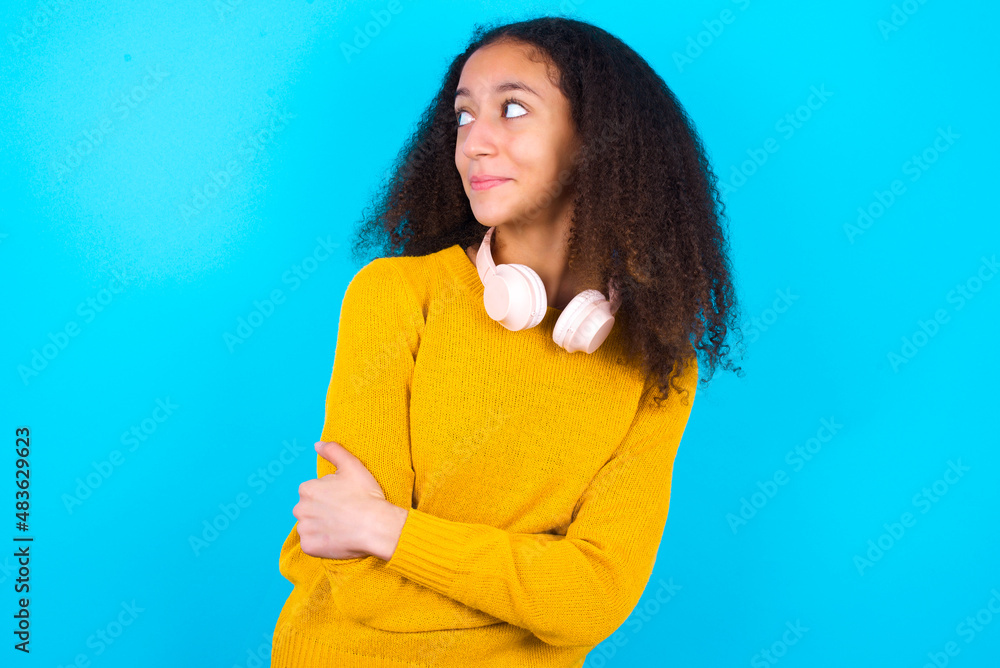 Pleased African teenager girl wearing yellow sweater over blue background keeps hands crossed over chest looks happily aside Pleased African teenager girl wearing yellow sweater over blue background keeps hands crossed over chest looks happily aside