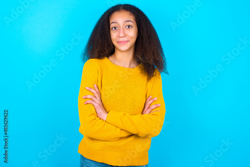 Self confident serious calm African teenager girl wearing yellow sweater over blue background stands with arms folded. Shows professional vibe stands in assertive pose.