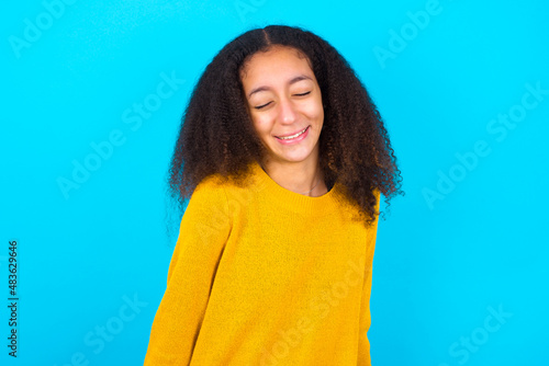 Positive African teenager girl wearing yellow sweater over blue background with overjoyed expression closes eyes and laughs shows white perfect teeth