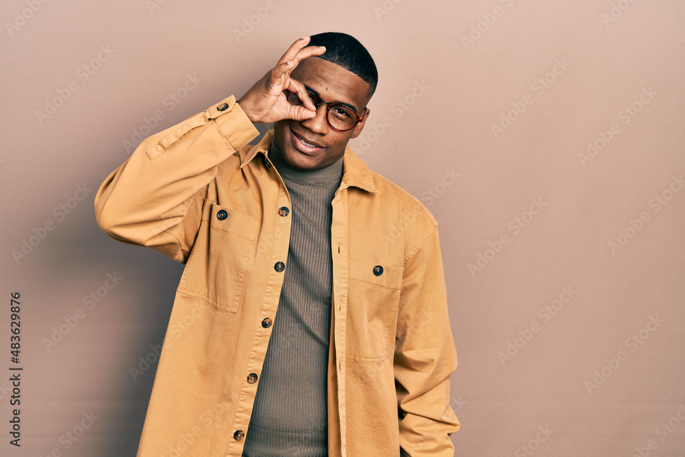 Young black man wearing casual clothes and glasses smiling happy doing ok sign with hand on eye looking through fingers Young black man wearing casual clothes and glasses smiling happy doing ok sign with hand on eye looking through fingers