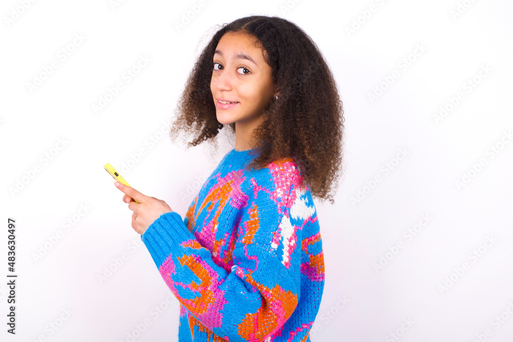 Rear view photo portrait of African teenager girl wearing colorful sweater over white background using smartphone smiling Rear view photo portrait of African teenager girl wearing colorful sweater over white background using smartphone smiling