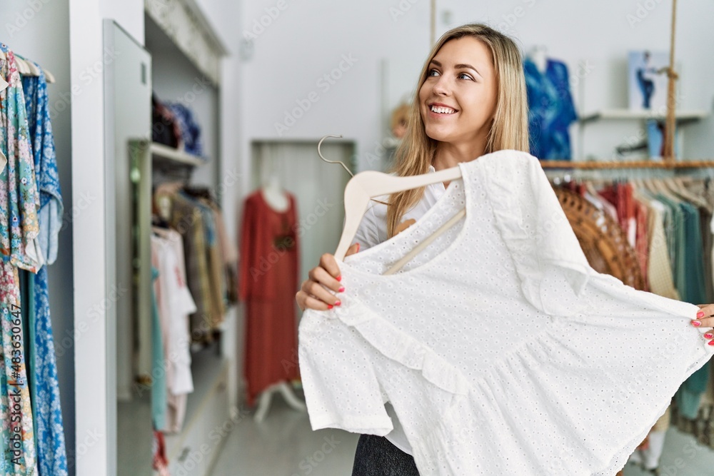 Young caucasian customer woman smiling happy holding hanger with clothes at clothing store. Young caucasian customer woman smiling happy holding hanger with clothes at clothing store.