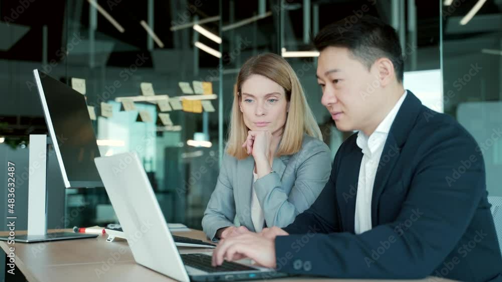 happy coworkers discuss project strategy by looking and pointing at laptop pc computer monitor screen. multiethnic business team in the office. confident mature asian man explaining young female happy coworkers discuss project strategy by looking and pointing at laptop pc computer monitor screen. multiethnic business team in the office. confident mature asian man explaining young female