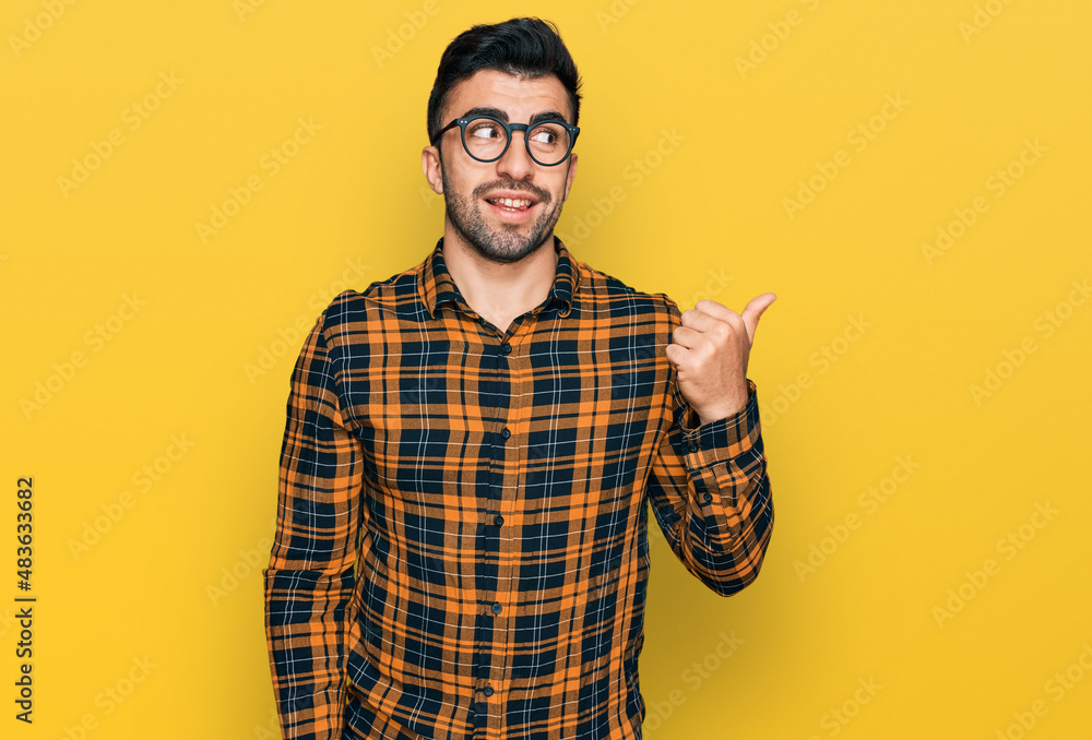 Hispanic man with beard wearing casual clothes and glasses smiling with happy face looking and pointing to the side with thumb up. Hispanic man with beard wearing casual clothes and glasses smiling with happy face looking and pointing to the side with thumb up.