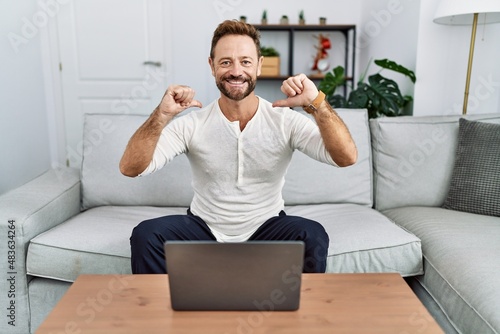 Middle age man using laptop at home looking confident with smile on face, pointing oneself with fingers proud and happy.