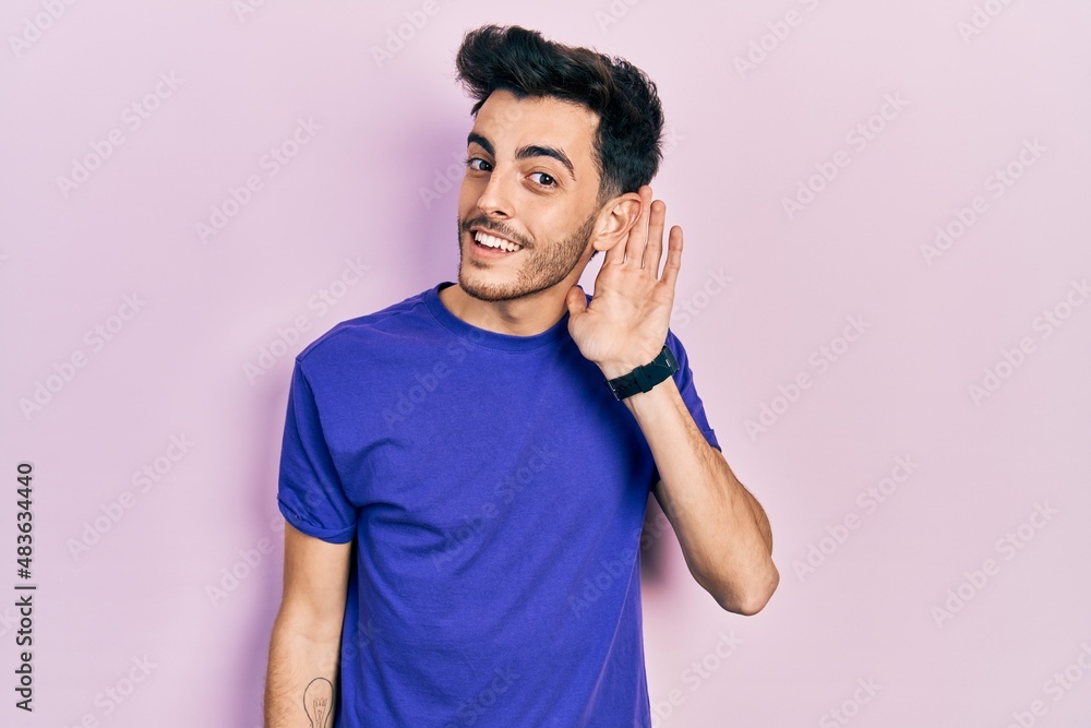 Young hispanic man wearing casual t shirt smiling with hand over ear listening an hearing to rumor or gossip. deafness concept. Young hispanic man wearing casual t shirt smiling with hand over ear listening an hearing to rumor or gossip. deafness concept.
