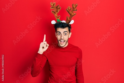 Young hispanic man wearing cute christmas reindeer horns pointing finger up with successful idea. exited and happy. number one.