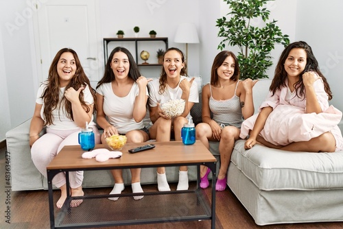 Group of women celebrating pajamas party at home pointing thumb up to the side smiling happy with open mouth