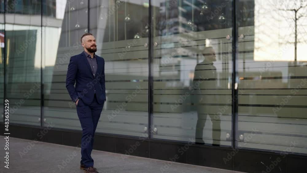 a moustached and bearded man in a blue suit has hands in pockets and is walking along the glass wall. the general plan a moustached and bearded man in a blue suit has hands in pockets and is walking along the glass wall. the general plan