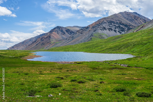 Colorful summer landscape with beautiful lake in sunlit green mountain valley among rocks and high mountain ridge under blue sky. Awesome scenery with alpine lake among greenery in sunlight.