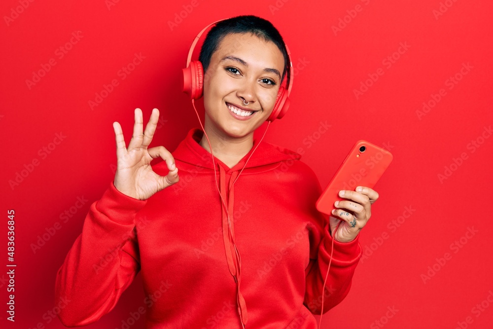 Beautiful hispanic woman with short hair using smartphone wearing headphones doing ok sign with fingers, smiling friendly gesturing excellent symbol Beautiful hispanic woman with short hair using smartphone wearing headphones doing ok sign with fingers, smiling friendly gesturing excellent symbol