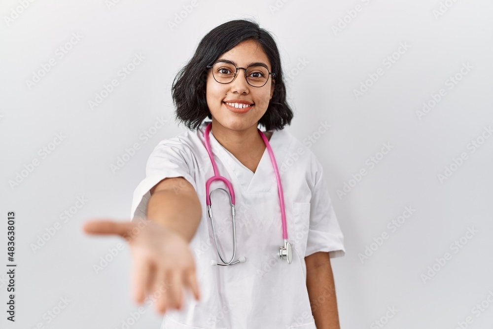 Young hispanic doctor woman wearing stethoscope over isolated background smiling friendly offering handshake as greeting and welcoming. successful business. Young hispanic doctor woman wearing stethoscope over isolated background smiling friendly offering handshake as greeting and welcoming. successful business.