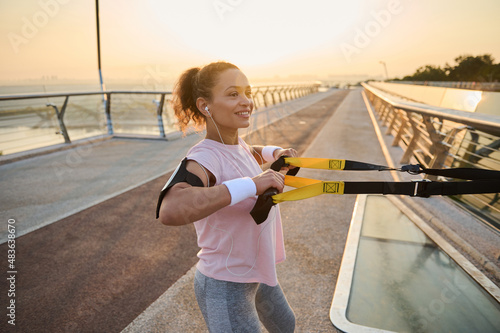Confident determined middle aged female athlete exercising with suspension straps, enjoying bodyweight training outdoor in the early morning at sunrise. Healthy habits and active lifestyle concept photo