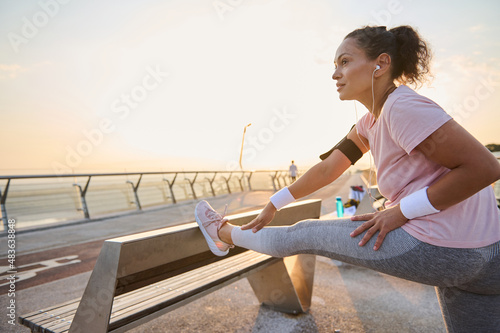 Female athlete, sportswoman in pink t-shirt stretching legs before morning jog. Sportswoman working out outdoor, standing on a city bridge and flexing her legs muscles on a wooden bench at sunrise